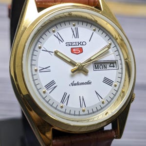 May include: A gold-toned Seiko 5 automatic wristwatch with a white face, Roman numeral hour markers, and a day-date display. The watch features gold hands and a brown leather strap. The watch face displays the day as "MON 4".