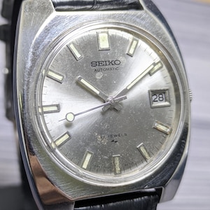 May include: A silver-toned Seiko Automatic wristwatch with a sunburst metallic dial, rectangular hour markers, and a date window at the three o'clock position. The watch features a brushed stainless steel case and a black textured leather strap.
