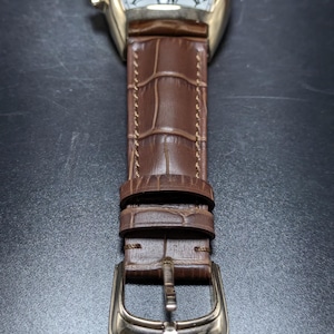 May include: A gold-tone wristwatch with a white dial featuring black Arabic numerals and chronograph sub-dials. The watch has a brown leather strap with a crocodile-embossed texture, brown stitching, and a gold-tone metal pin buckle closure.