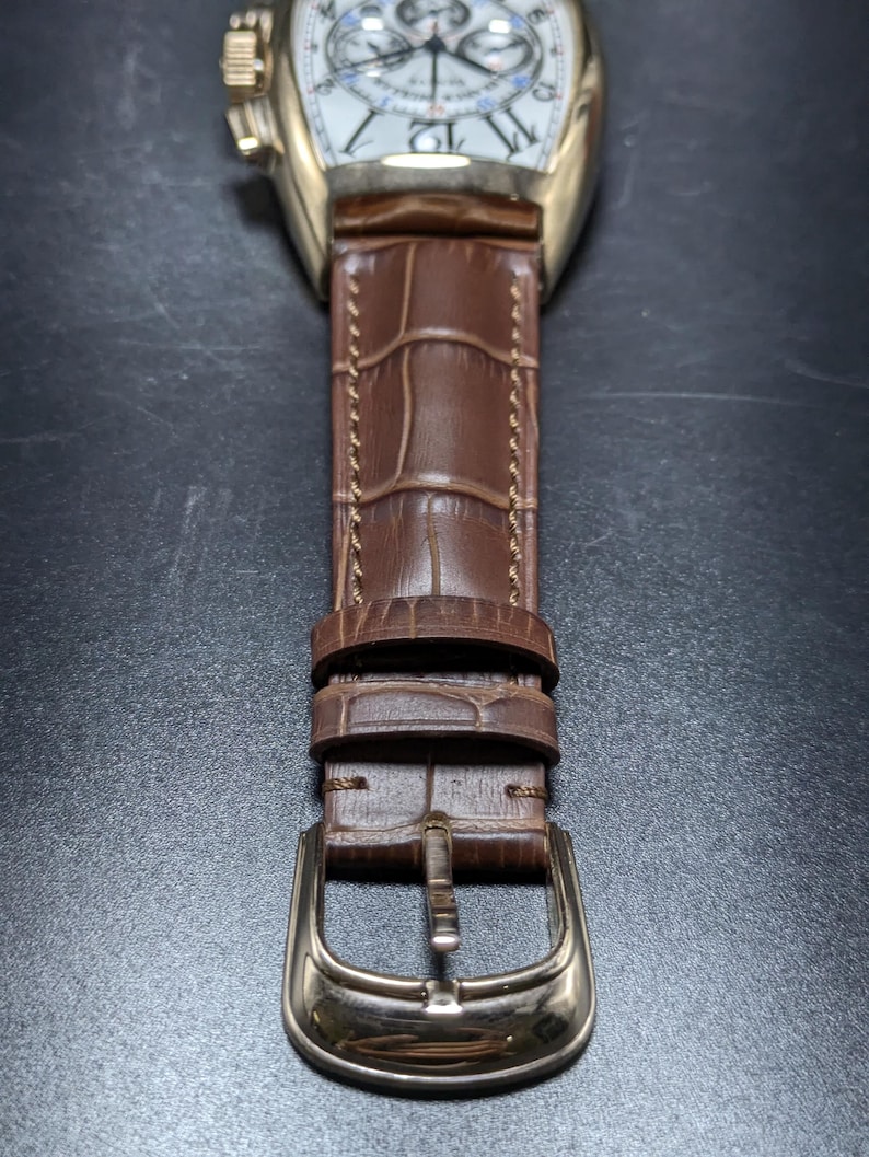 May include: A gold-tone wristwatch with a white dial featuring black Arabic numerals and chronograph sub-dials. The watch has a brown leather strap with a crocodile-embossed texture, brown stitching, and a gold-tone metal pin buckle closure.