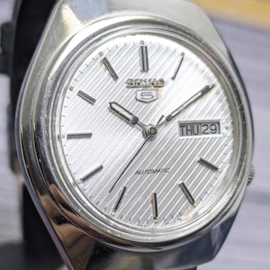 May include: A silver-toned Seiko 5 automatic wristwatch with a textured, diagonal-striped dial. The watch features silver hour markers, silver hands, and a rectangular day-date window displaying THU 29. The case is polished stainless steel.