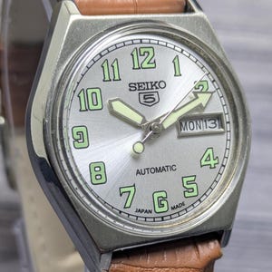 Peut inclure: Une montre-bracelet automatique Seiko 5 argentée dotée d'un cadran soleillé avec des chiffres arabes verts luminescents, des aiguilles luminescentes et un guichet jour-date à trois heures. La montre est fixée à un bracelet en cuir marron texturé.