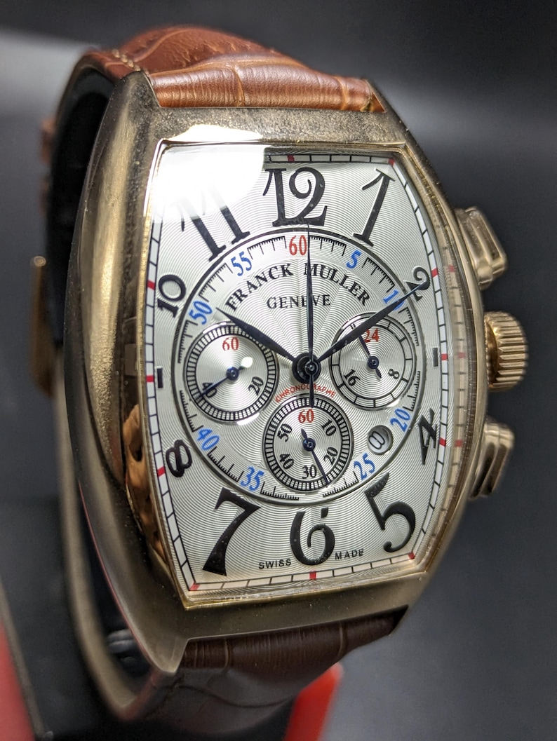 May include: A Franck Muller Geneve chronograph watch with a tonneau-shaped gold case, a white guilloch&eacute; dial featuring black Arabic numerals, three sub-dials, and a brown leather strap with a textured crocodile pattern and a gold-tone buckle.