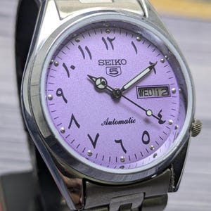 May include: A silver-toned Seiko 5 automatic wristwatch with a lavender dial. The watch face features Arabic numerals, a day-date display, and luminous hands. The watch has a stainless steel band.