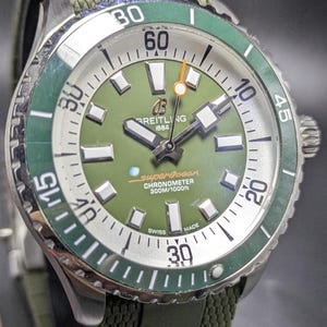 Könnte beinhalten: Eine Breitling Superocean Armbanduhr mit grünem Zifferblatt und Lünette. Die Uhr hat ein silbernes Gehäuse, ein grünes Stoffarmband und den Text "Breitling 1884" auf dem Zifferblatt. Die Uhr ist wasserdicht bis 300 Meter.