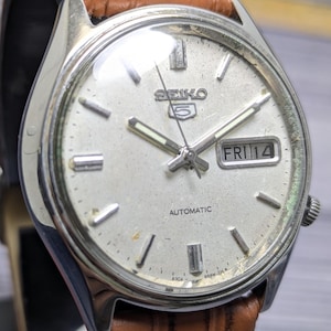 May include: A silver-toned Seiko 5 automatic wristwatch with a textured off-white dial, silver baton hour markers, and luminous hands. The watch features a day-date window at the three o'clock position displaying FRI 14 and a brown leather strap.
