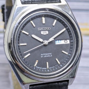May include: A vintage Seiko 5 automatic wristwatch with a black dial. The watch features silver-tone hands and markers, a day-date display, and a stainless steel case. The watch has a black leather strap.