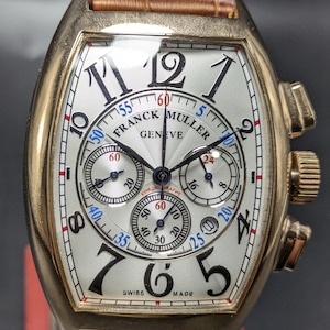 May include: A Franck Muller Geneve chronograph wristwatch with a tonneau-shaped gold case, a white guilloche dial featuring black stylized Arabic numerals, three sub-dials, and a brown leather strap with a textured crocodile-like pattern.