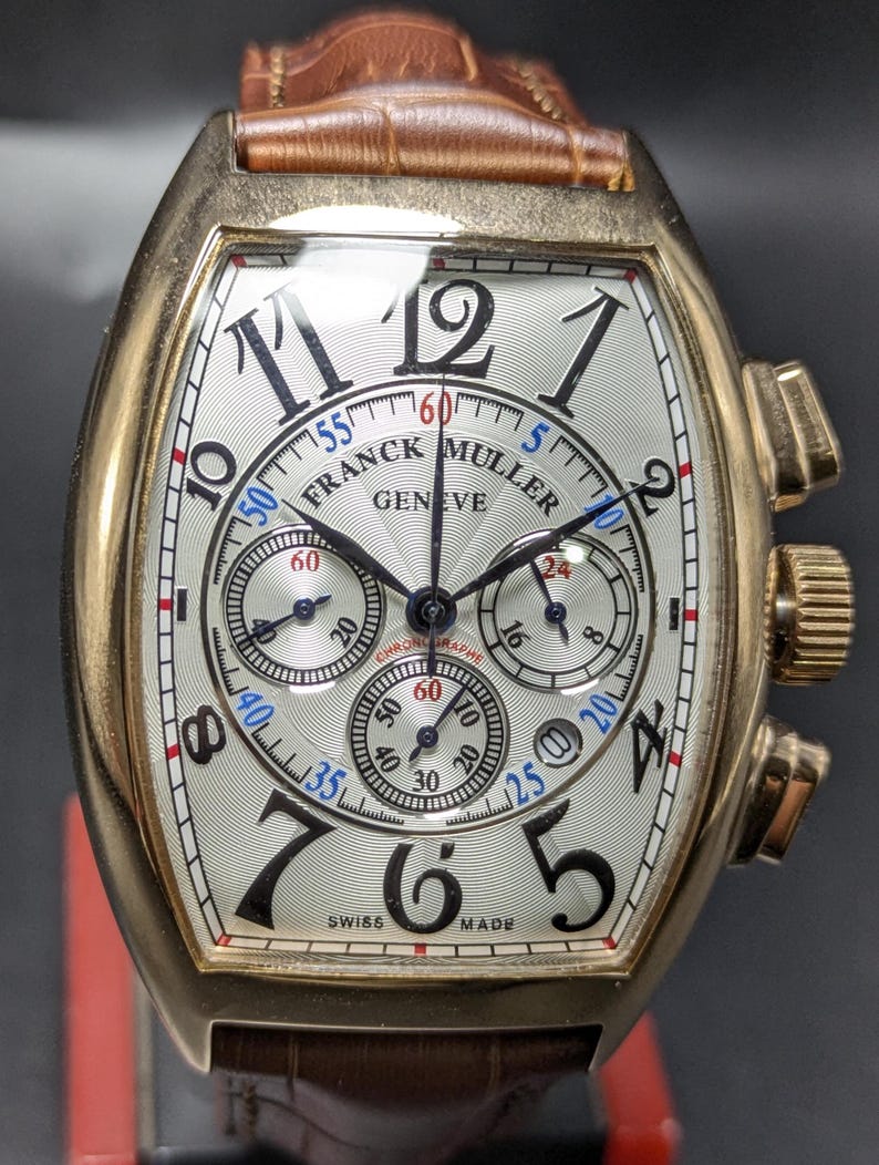 May include: A Franck Muller Geneve chronograph wristwatch with a tonneau-shaped gold case, a white guilloche dial featuring black stylized Arabic numerals, three sub-dials, and a brown leather strap with a textured crocodile-like pattern.
