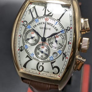 May include: A gold-toned Franck Muller Geneve chronograph watch with a tonneau-shaped case, a silver guilloch&eacute; dial featuring black Arabic numerals, three sub-dials, and a brown leather strap with a textured, crocodile-embossed pattern.