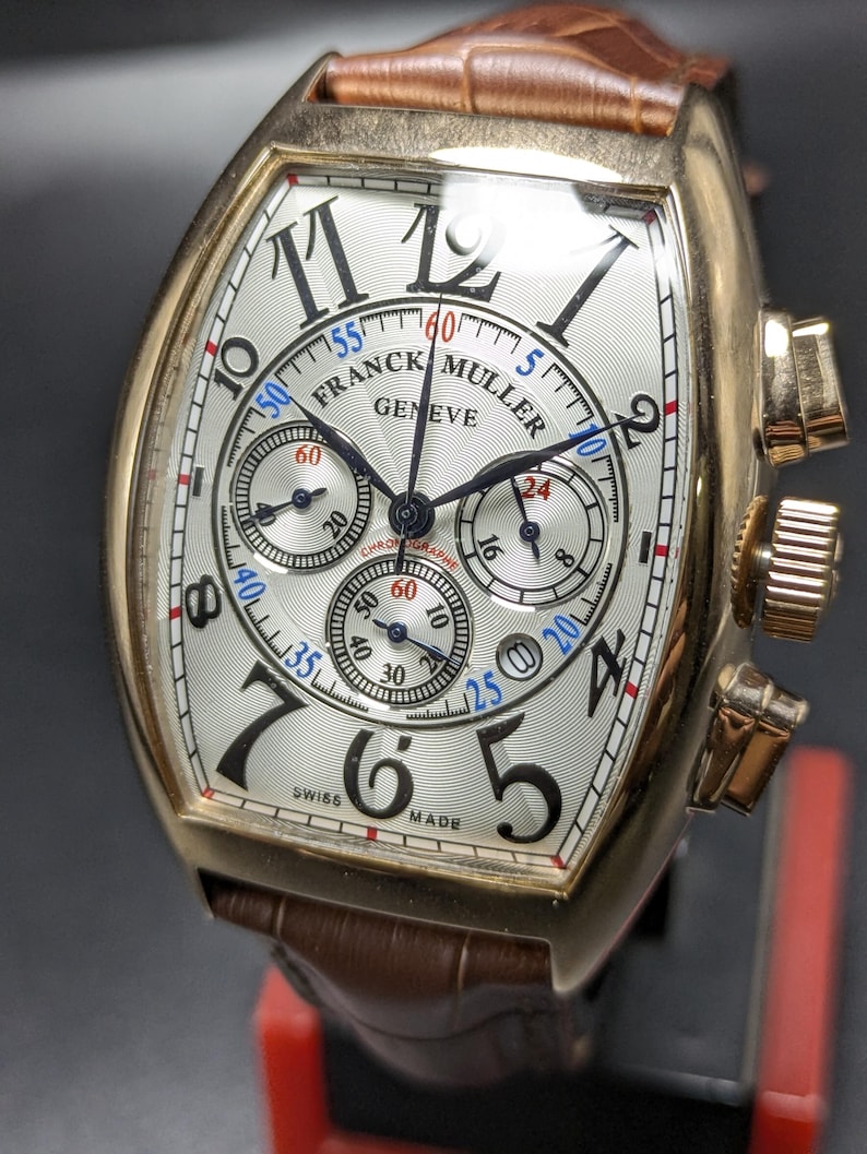 May include: A gold-toned Franck Muller Geneve chronograph watch with a tonneau-shaped case, a silver guilloch&eacute; dial featuring black Arabic numerals, three sub-dials, and a brown leather strap with a textured, crocodile-embossed pattern.