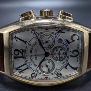 May include: A gold-toned tonneau-shaped chronograph watch with a silver dial, black Arabic numerals, and three sub-dials. The watch features a brown leather strap, a crown, and two pushers at the top. The dial displays the text Franck Muller Geneve.