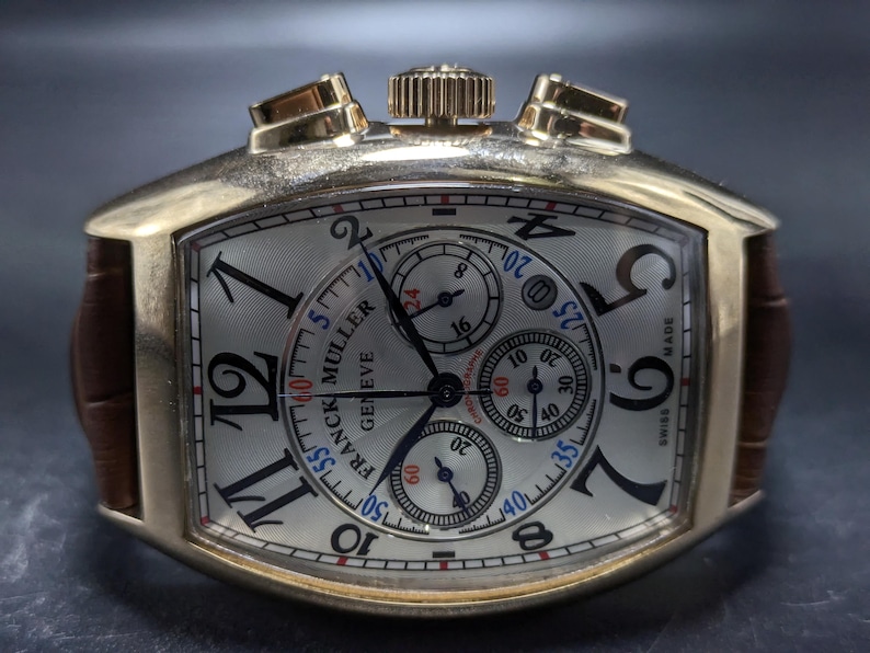 May include: A gold-toned tonneau-shaped chronograph watch with a silver dial, black Arabic numerals, and three sub-dials. The watch features a brown leather strap, a crown, and two pushers at the top. The dial displays the text Franck Muller Geneve.