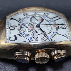 May include: A gold-toned Franck Muller Geneve chronograph wristwatch with a white dial, black Arabic numerals, and three sub-dials. The watch features a brown leather strap with a textured finish, a fluted crown, and two side pushers.