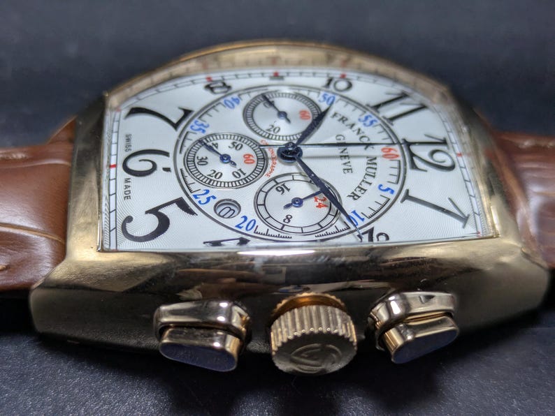 May include: A gold-toned Franck Muller Geneve chronograph wristwatch with a white dial, black Arabic numerals, and three sub-dials. The watch features a brown leather strap with a textured finish, a fluted crown, and two side pushers.