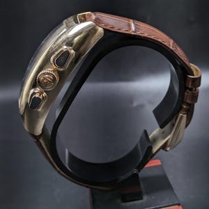 May include: A gold-tone wristwatch with a brown leather strap featuring a crocodile-embossed texture. The watch case has a polished finish, two side pushers, and a central crown with an engraved logo. The strap includes a metal buckle closure.