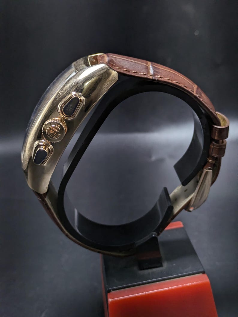 May include: A gold-tone wristwatch with a brown leather strap featuring a crocodile-embossed texture. The watch case has a polished finish, two side pushers, and a central crown with an engraved logo. The strap includes a metal buckle closure.