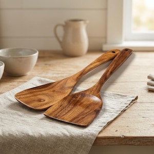 May include: Two wooden kitchen spatulas with long handles and a natural wood grain finish. One spatula is flat and wide, while the other is more narrow and angled. They rest on a linen cloth.