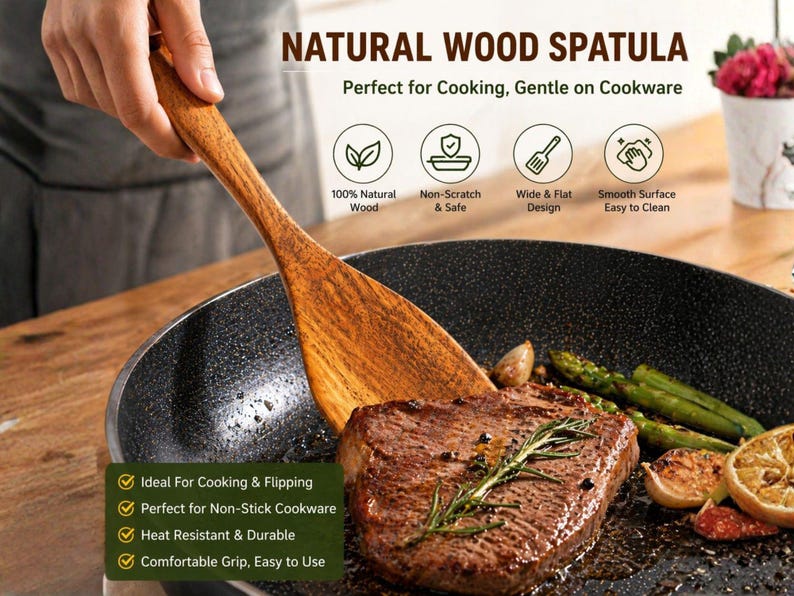 May include: A wooden spatula is shown in use, flipping a steak in a non-stick pan. The spatula is made of light brown wood and has a wide, flat design. The pan contains cooked steak, asparagus, and lemon slices.
