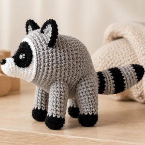 May include: A crocheted raccoon stuffed animal featuring a grey body, black and white facial markings, black ears, and black paws. The raccoon has a long, striped tail with alternating black and grey bands and shiny black plastic eyes.