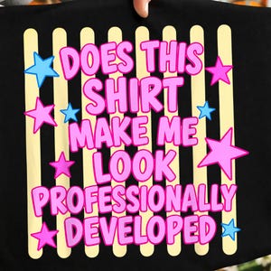 May include: A black t-shirt featuring a graphic print of vertical pale yellow stripes. The text reads "DOES THIS SHIRT MAKE ME LOOK PROFESSIONALLY DEVELOPED" in pink bubble letters, surrounded by scattered light blue and pink five-pointed stars.