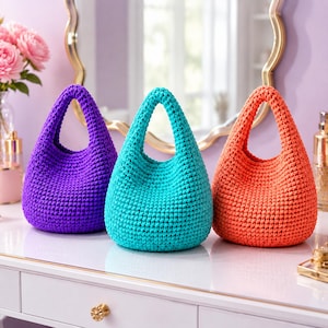 Puede incluir: Tres bolsos pequeños y redondeados hechos de hilo grueso y texturizado en colores sólidos vibrantes. El bolso de la izquierda es morado, el central es turquesa y el de la derecha es naranja brillante. Cada bolso tiene un asa integrada.