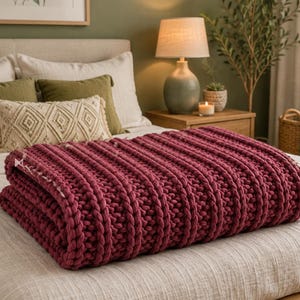 Op de afbeelding: Een dikke, grof gebreide plaid in een diepe bordeauxrode kleur met een opvallende verticale ribbelstructuur. De deken is netjes opgevouwen en over een lichtgekleurde sprei gelegd, wat de zware garenconstructie en het patroon goed laat zien.