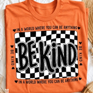 May include: An orange short-sleeved t-shirt featuring a black and white checkered rectangle graphic. The text "BE KIND" is printed in large black letters over the pattern, surrounded by the phrase "IN A WORLD WHERE YOU CAN BE ANYTHING".