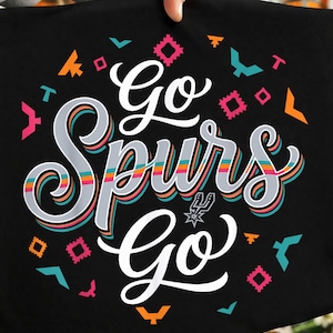 Puede incluir: Camiseta negra con la frase "Go Spurs Go" en una fuente estilizada y colorida. El diseño incluye formas geométricas en naranja, rosa y turquesa, con el logotipo de los Spurs. La camiseta está sujeta por una persona.