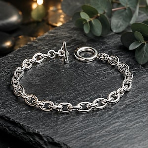 May include: A silver-toned chain bracelet featuring a series of interconnected oval links. The bracelet is secured with a toggle clasp, consisting of a T-bar and a circular ring, which serves as both a functional closure and a decorative element.