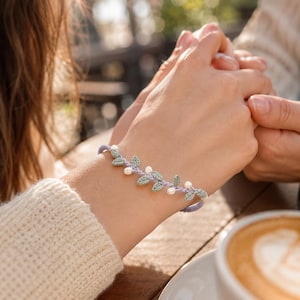 Op de afbeelding: Een delicate armband met een lichtpaars gevlochten koord, versierd met kleine, saliegroene gehaakte blaadjes en piepkleine witte bloemaccenten. Het ontwerp bootst een rankachtig patroon na dat om de pols wikkelt en toont fijn handwerk.