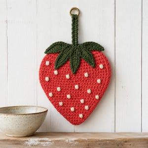 Op de afbeelding: Een aardbeivormige gehaakte pannenlap in rood met kleine witte textuurstippen en een donkergroene bladeren bovenkant. Het item heeft een stevige donkergroene gehaakte lus aan de bovenkant, bevestigd aan een goudkleurige metalen ring om op te hangen.