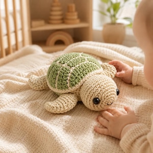 May include: A crochet turtle toy featuring a textured sage green shell with cream-colored borders and a light beige body. The stuffed animal has two large, round, black plastic eyes and a simple stitched smile, resting on a soft, cream-colored blanket.