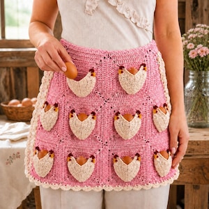 May include: A pink crocheted apron featuring a grid of cream-colored chicken-shaped pockets, each holding a brown egg. The apron has a scalloped cream border and is worn over a white top. The design includes small red and yellow accents on the chickens.