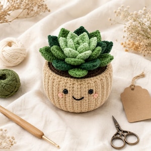 May include: A crocheted succulent plant in a beige pot with a stitched smiling face, featuring layered green leaves in varying shades. The pot has two black bead eyes and pink embroidered cheeks, crafted with textured yarn stitches throughout.