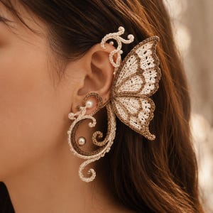 May include: An ear cuff featuring a crocheted butterfly wing design in cream and brown thread. The piece includes intricate swirling patterns that wrap around the ear, accented with small white pearls integrated into the crocheted structure.
