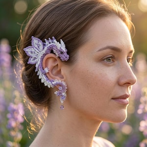 Può includere: Un ear cuff all'uncinetto a forma di drago, realizzato con filato viola chiaro e bianco. Il design del drago avvolge l'orecchio esterno, con una piccola perla viola che pende dal fondo. La lavorazione mostra ali e squame molto dettagliate.