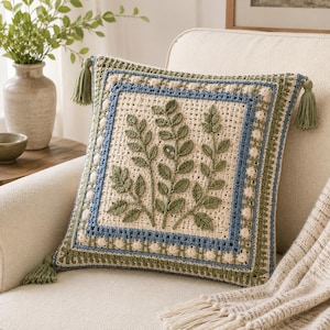 May include: A square crochet pillow featuring a cream-colored center with three raised green leaf motifs. The border consists of alternating bands of blue and green yarn with small cream bobbles. Four green tassels are attached to the corners.