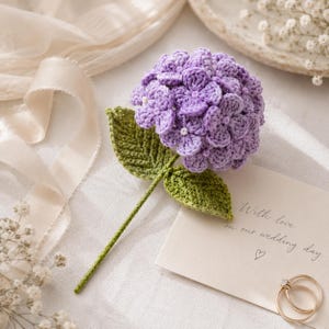 Puede incluir: Una flor de hortensia morada tejida a ganchillo con un tallo verde y dos hojas verdes. La cabeza de la flor se compone de múltiples pétalos pequeños y detallados con diminutos centros blancos. El tallo es fino y recto, desde la base.