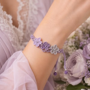 May include: A delicate crochet bracelet featuring three interconnected flower motifs in varying shades of purple and lavender. The floral design is accented with small silver-toned beads at the center of each flower and at the ends of the adjustable cord.