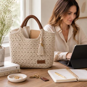 Könnte beinhalten: Eine beige gehäkelte Tragetasche mit zwei braunen lederartigen Griffen und einem dekorativen Quastenanhänger. Die Tasche verfügt über ein rechteckiges braunes Etikett mit dem eingeprägten Wort Handmade auf der Vorderseite und ein strukturiertes Muster.