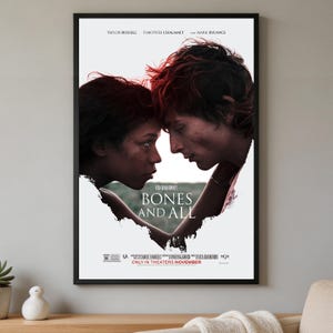 May include: A framed movie poster for Bones and All featuring Taylor Russell and Timothée Chalamet facing each other. The title Bones and All is printed in white text in the center. The background is white with a torn paper effect around the actors.
