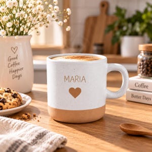 Op de afbeelding: Een witte keramische mok met een gespikkelde afwerking en een beige basis. De voorzijde van de mok toont de naam MARIA in bruine letters boven een bruin harticoon. De mok heeft een dik, rond handvat en bevat een lichtbruine drank.