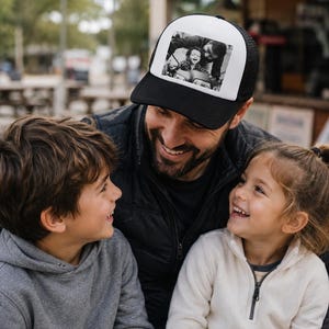 Puede incluir: Una gorra de estilo camionero en blanco y negro con una impresión fotográfica rectangular personalizada en el panel frontal. La gorra tiene una visera negra sólida, un panel frontal blanco y una parte trasera de malla negra con cierre ajustable.