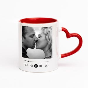 Può includere: Una tazza in ceramica bianca con interno rosso e manico rosso a forma di cuore. La parte anteriore presenta una fotografia in bianco e nero di una coppia che si bacia, posizionata sopra un'interfaccia di lettore musicale digitale.