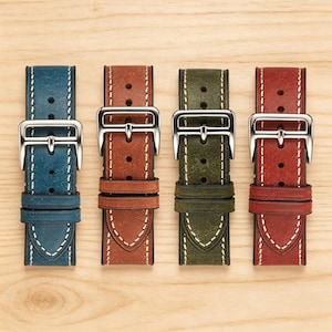 May include: Four leather watch straps in blue, brown, olive green, and red. Each strap has a silver buckle and white stitching. The straps are arranged vertically on a light wood surface.