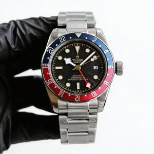 May include: A stainless steel wristwatch with a black dial, a red and blue bezel, and a silver band. The watch face displays the brand name "TUDOR GENEVE" and other text. The watch has a date display at the 3 o'clock position.