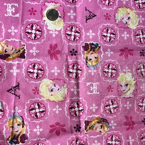 May include: Pink fabric with a repeating pattern of princesses from the movie Frozen. The princesses are wearing blue and purple dresses and have blonde hair. The background is a pink and white geometric pattern.