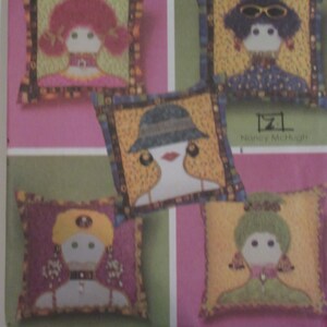 May include: Four colourful pillow designs featuring stylised faces with different hair colours, hats, and accessories. The pillows are all square and have a patterned border. The text "Nancy McHugh" is visible in the bottom right corner.