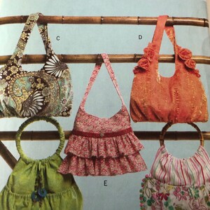 Puede incluir: Cinco estilos diferentes de bolsos de tela colgados de un estante de bambú. Los bolsos están hechos de varias telas y colores, incluyendo estampados florales, rayas y colores sólidos. Los bolsos tienen diferentes tipos de asas, incluyendo correas, lazos y anillos.
